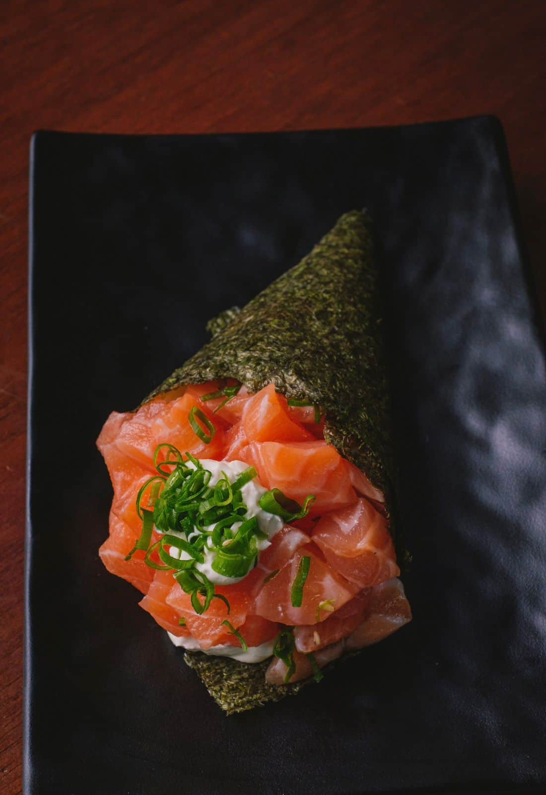 A hand roll sushi with diced salmon, green onions, and mayonnaise, wrapped in a sheet of nori seaweed, served on a black rectangular plate over a dark wooden surface.