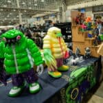 A convention booth displays colorful, large monster toys in jackets, with more monster figures and small sculptures arranged on a table and shelves. People are browsing the busy indoor event space in the background.
