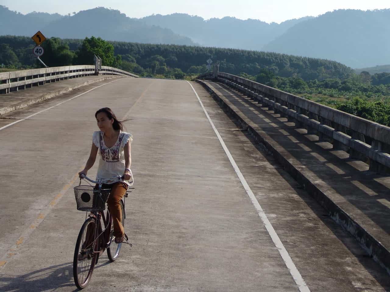 A woman rides a bicycle on an empty, sunlit road with concrete barriers, evoking scenes from Southeast Asian cinema, surrounded by green hills and distant mountains under a clear sky.