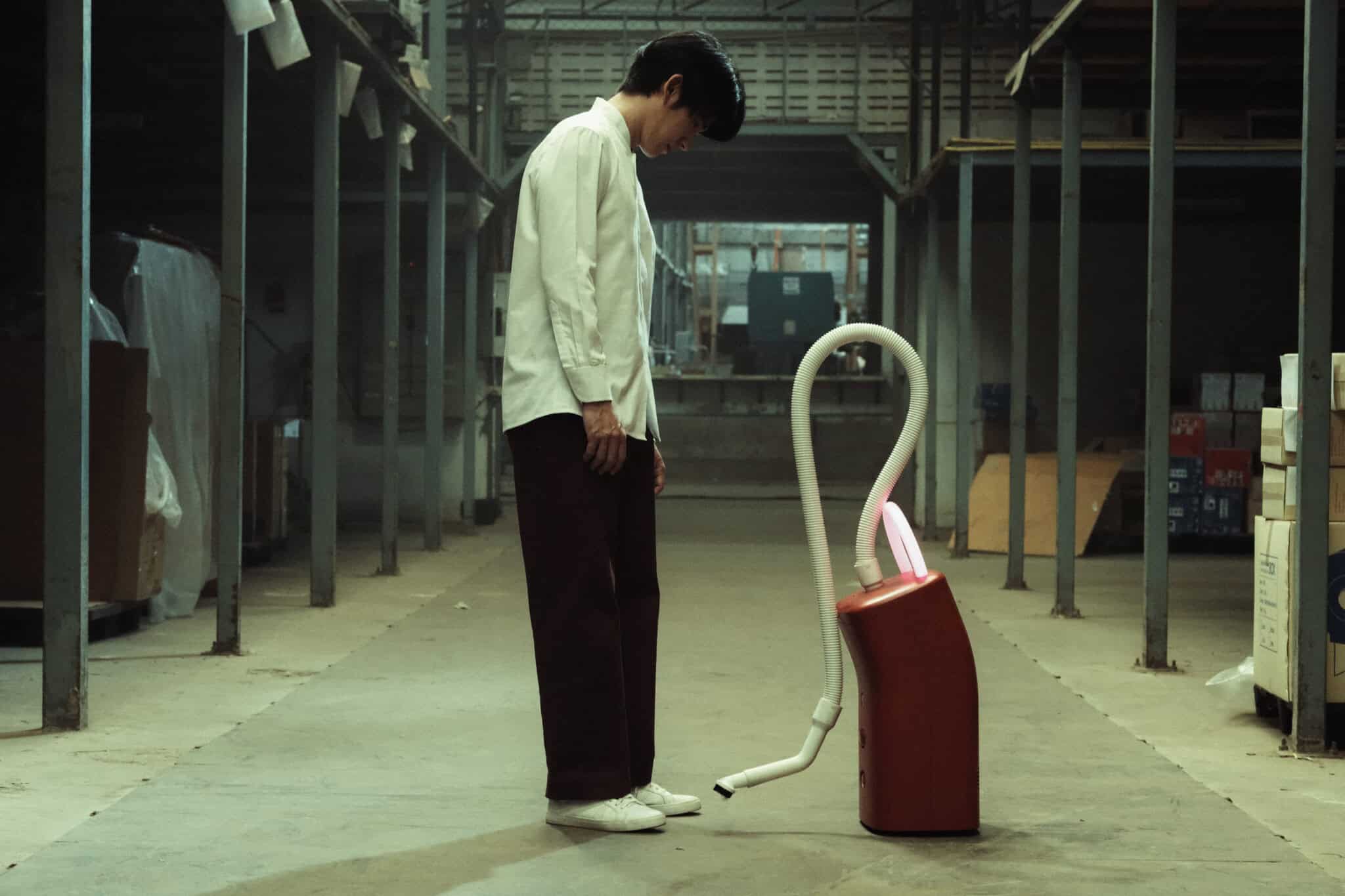 A person in a white shirt and dark pants stands in an industrial warehouse, facing a red vacuum cleaner with a white hose, creating a sense of interaction or contemplation.