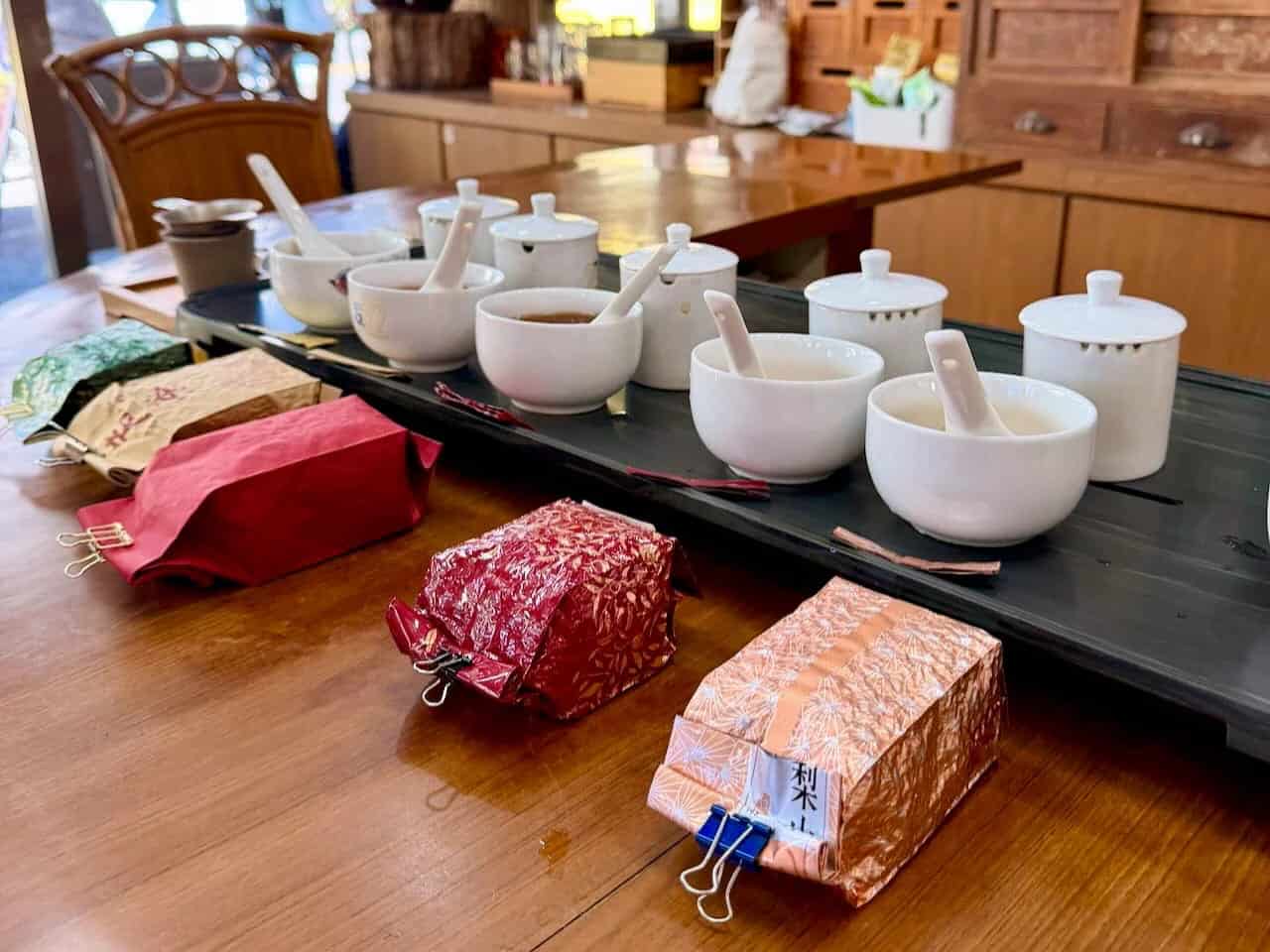 A wooden table displays several white ceramic tea cups with lids and spoons, alongside colorful, textured tea packages secured with binder clips, set in a cozy, traditional tea shop.