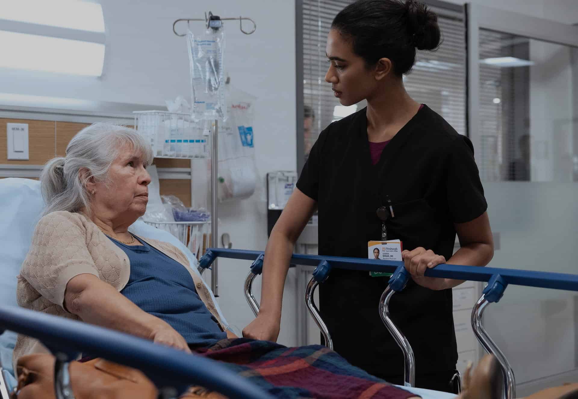 A nurse in black scrubs stands beside an elderly woman lying in a hospital bed, talking to her and holding the beds side rail. The patient looks concerned while they have a conversation in a hospital room.