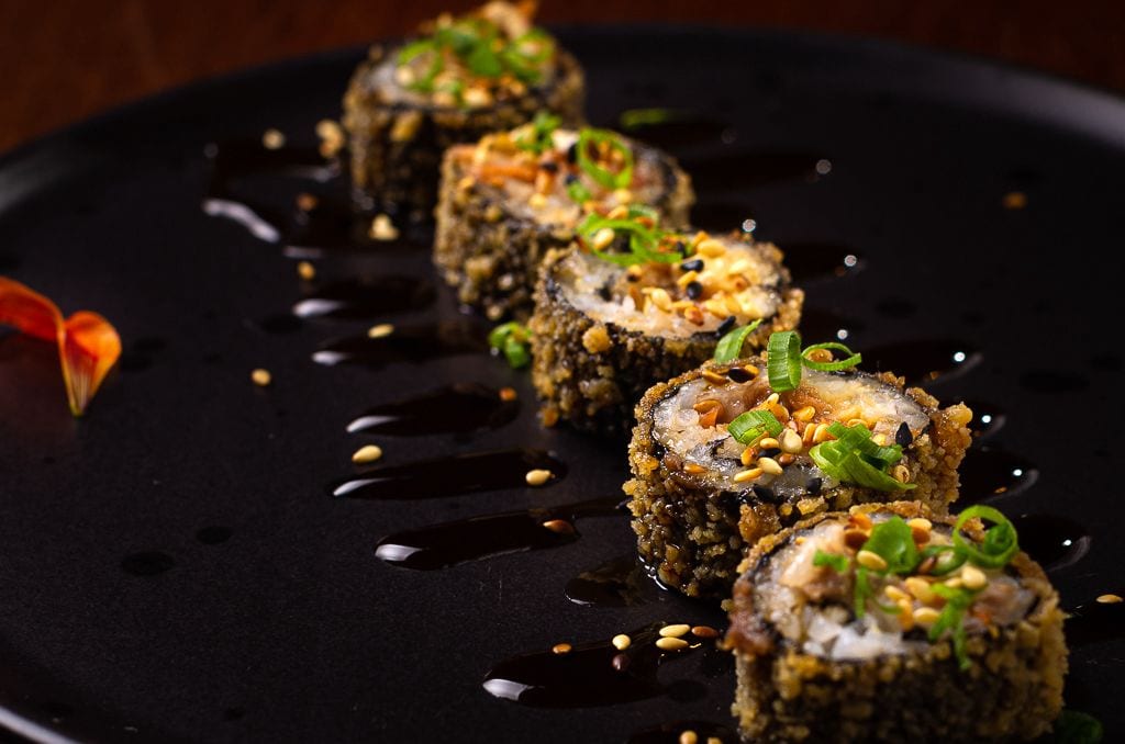Five pieces of deep-fried sushi rolls are arranged on a black plate, garnished with chopped green onions and sesame seeds, and drizzled with a dark sauce.