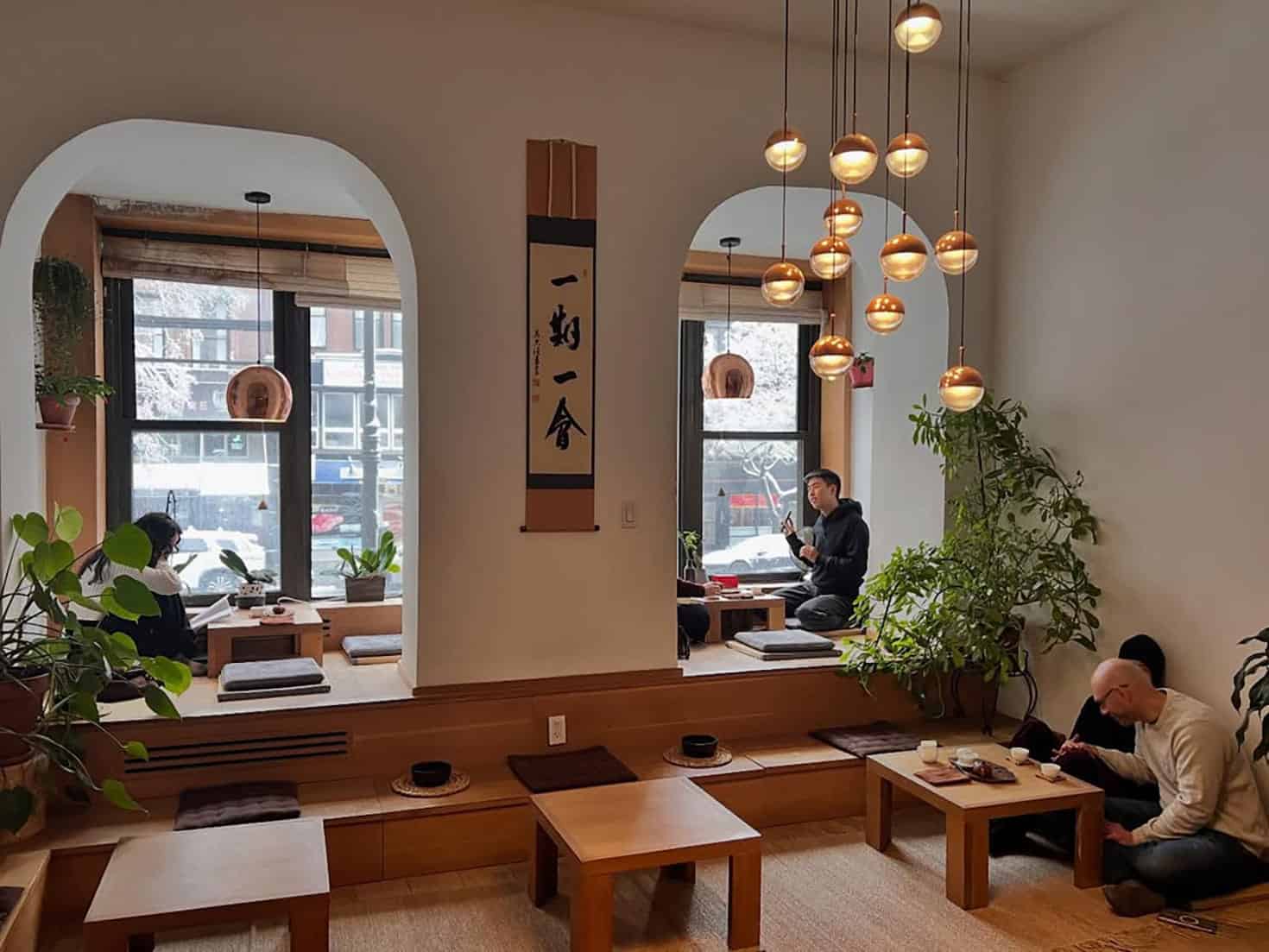 A cozy, minimalist café with low wooden tables, floor cushions, hanging pendant lights, lush green plants, and large windows. Two people sit near the windows, and another person sits alone at a table, all enjoying a peaceful atmosphere.