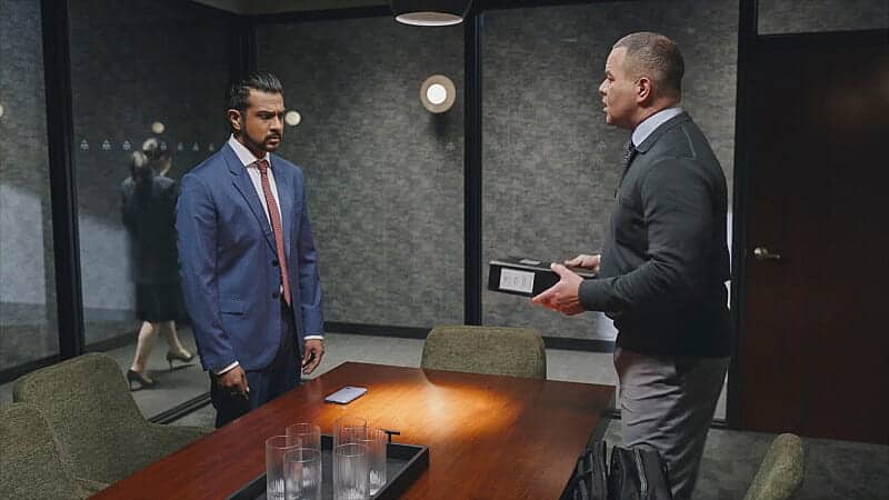 Two men are in a modern conference room having a serious conversation; one stands by the table in a blue suit, while the other holds a box. A woman is seen walking outside through the glass wall.