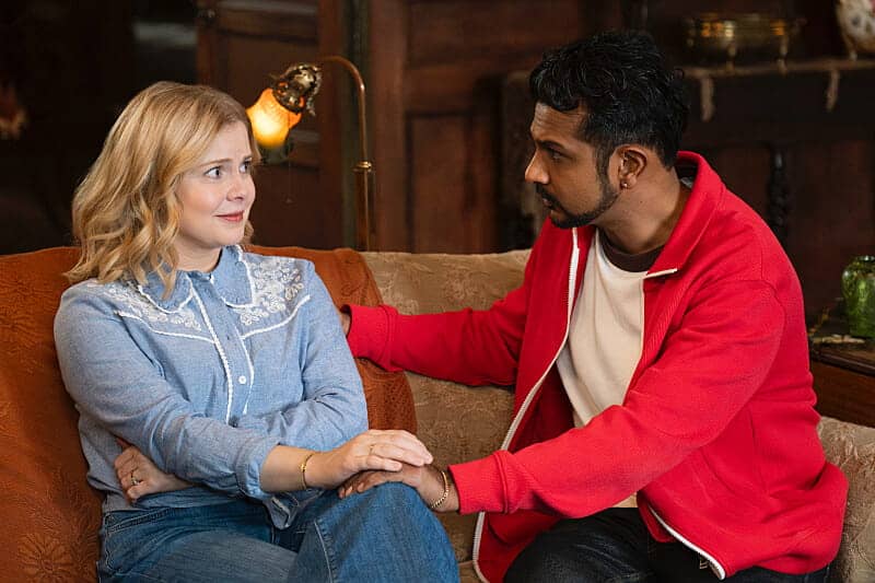 A woman and a man sit on a couch indoors. The man, wearing a red jacket, gently touches the womans arm, while she looks at him with a surprised or uncertain expression. They appear to be having a serious conversation.