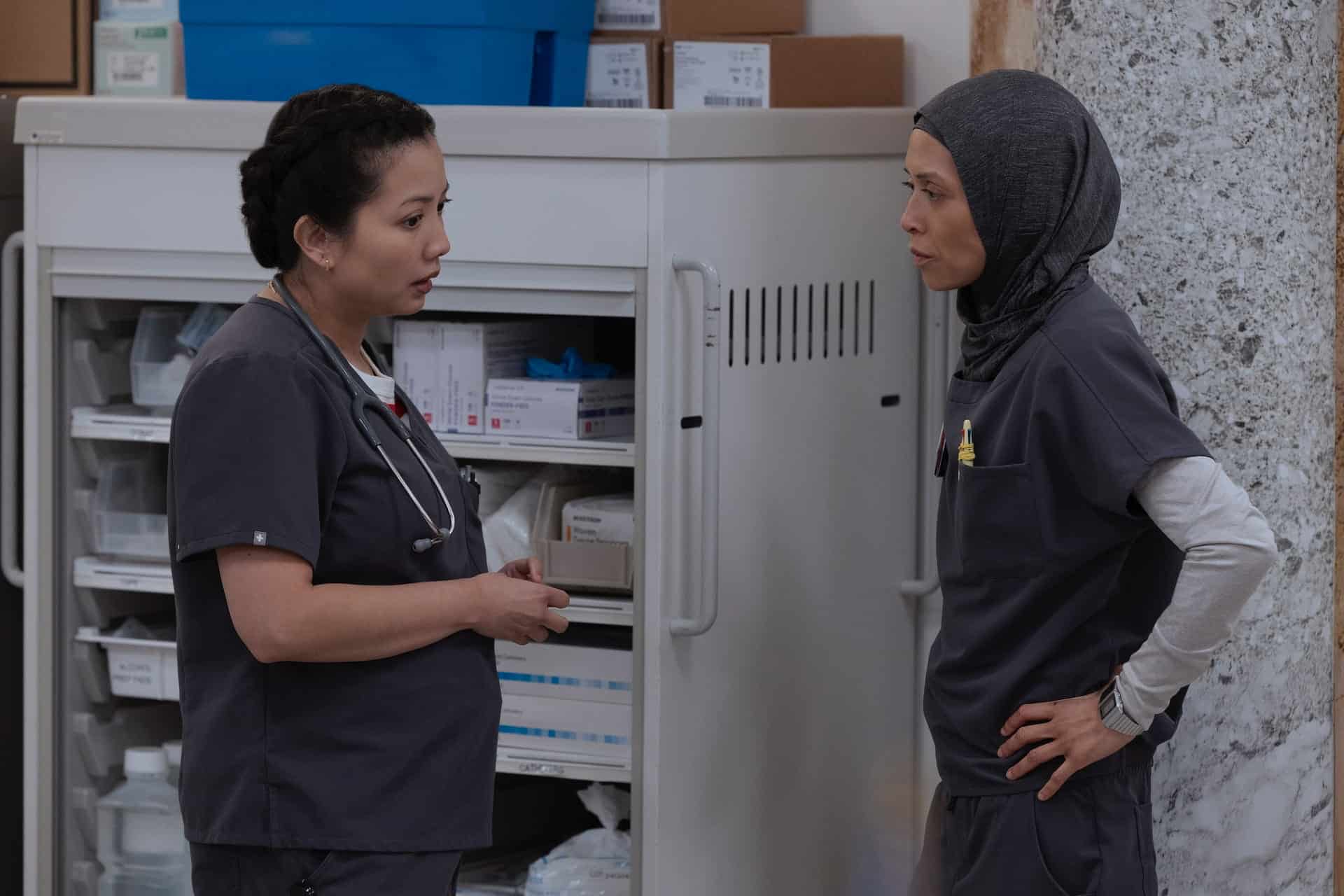 Two female healthcare professionals in gray scrubs stand and talk in front of a medical supply cart. One has a stethoscope around her neck, and the other wears a gray headscarf. They appear to be having a serious conversation.
