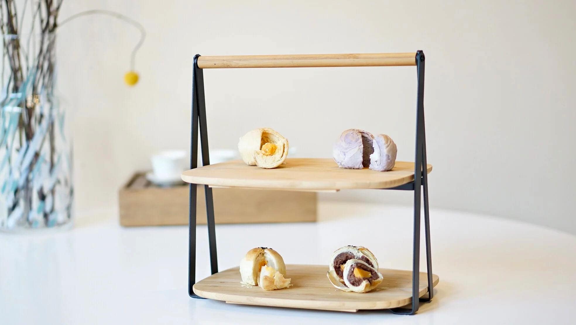 A two-tier wooden stand with four assorted pastries, two on each level, displayed on a white table. A blurred vase with branches and a teacup are in the background.