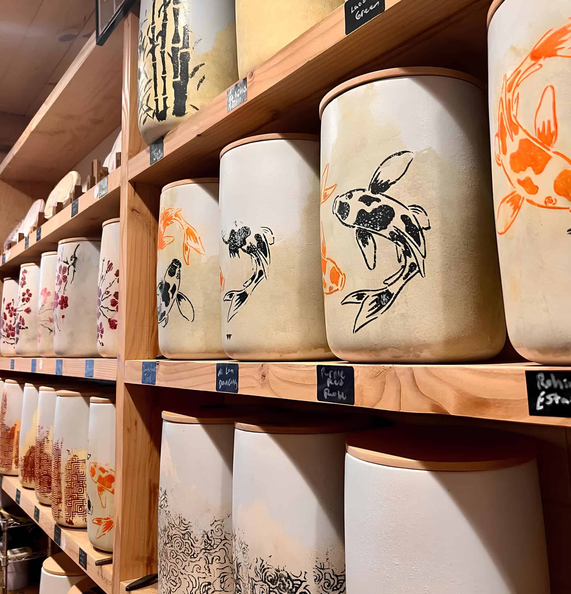 Large ceramic jars with hand-painted koi fish and other designs are neatly arranged on wooden shelves. Each jar is labeled with a small black sign, and the shelves are set against a wooden wall in a shop.
