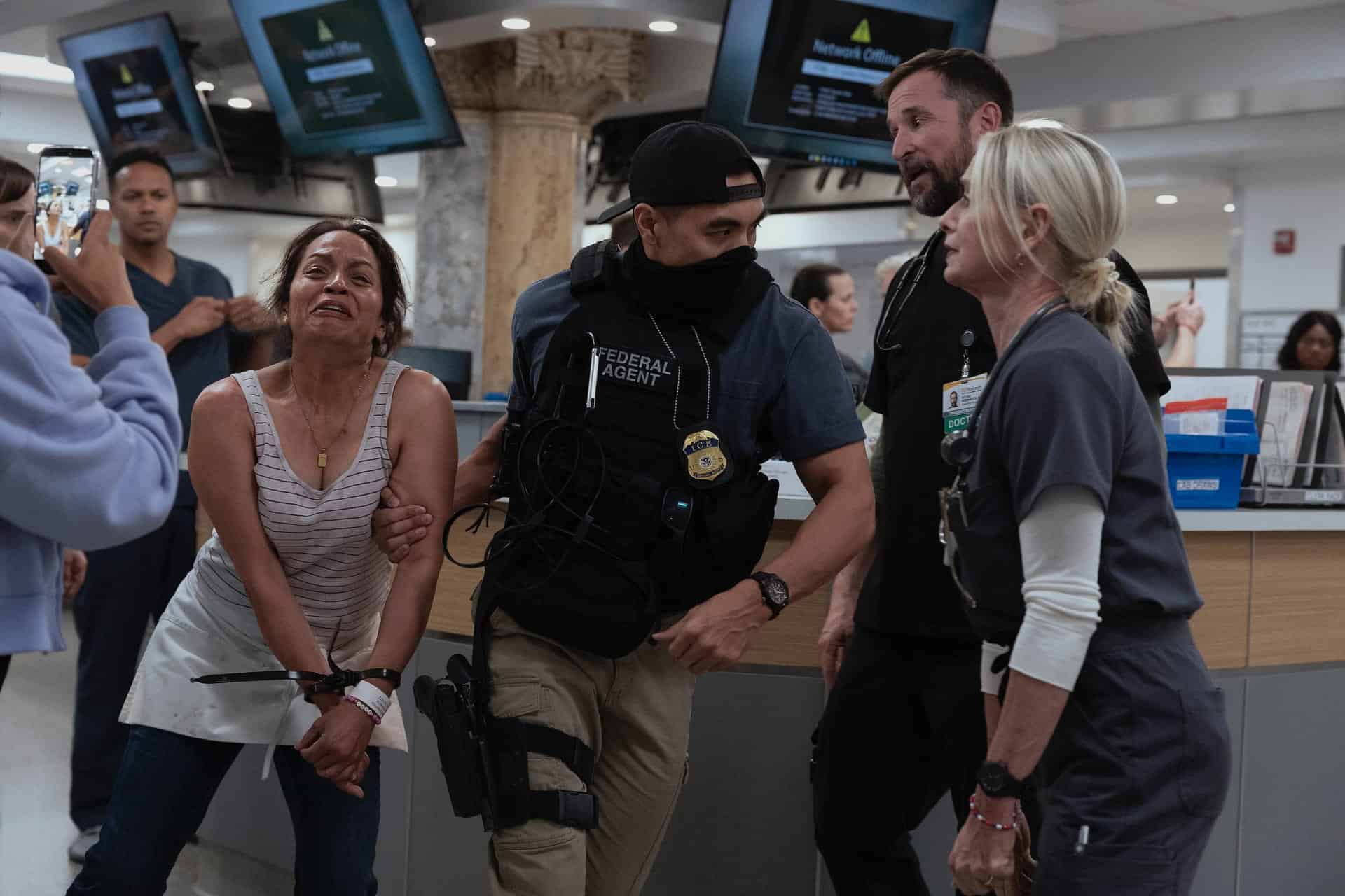 A distressed woman with her hands bound is escorted by a federal agent and hospital staff in a busy hospital setting, while another person films the scene with a phone.