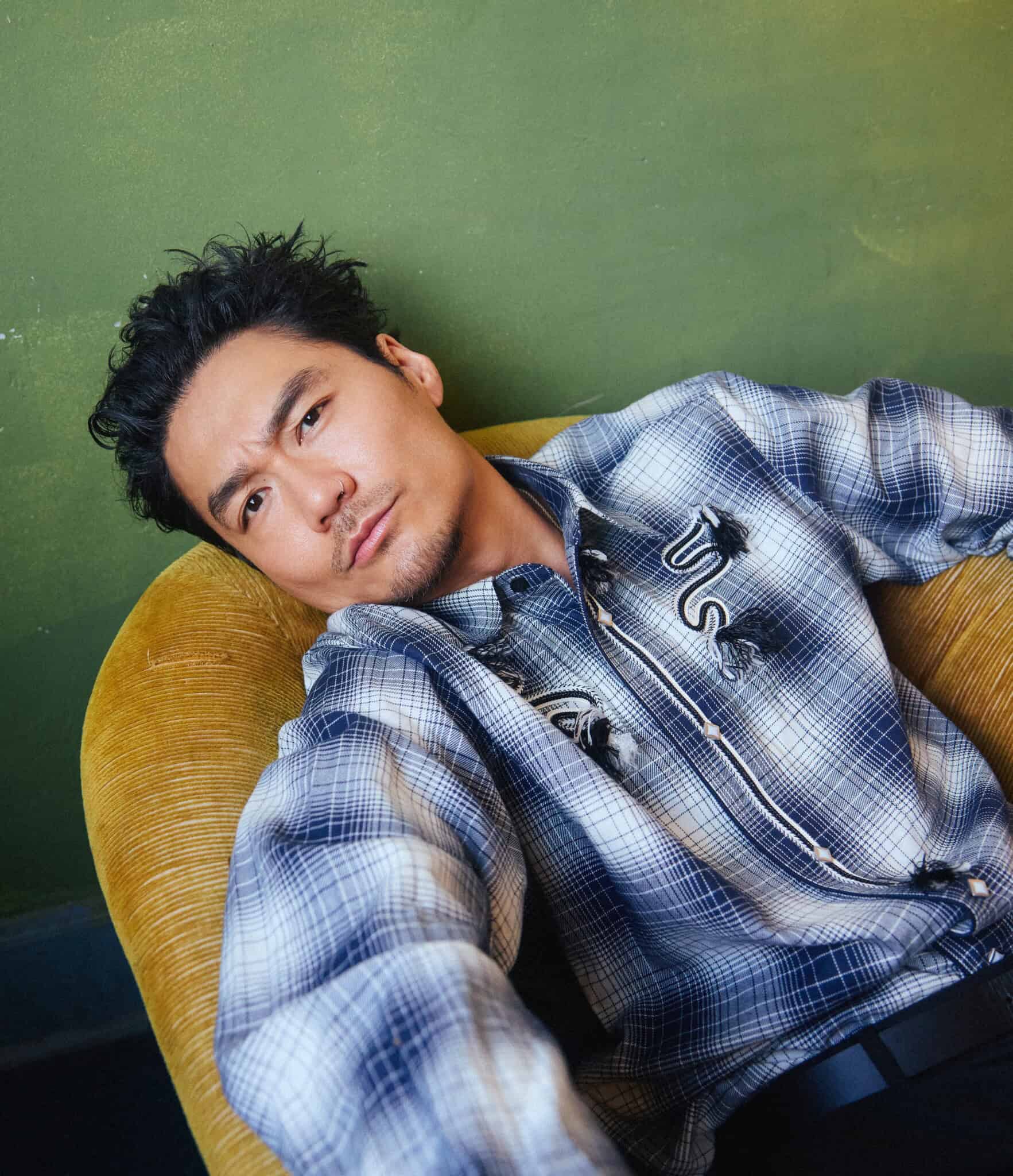 A man with short, styled black hair and slight facial hair reclines in a yellow chair, wearing a blue plaid shirt with embroidered details, against a green wall background.