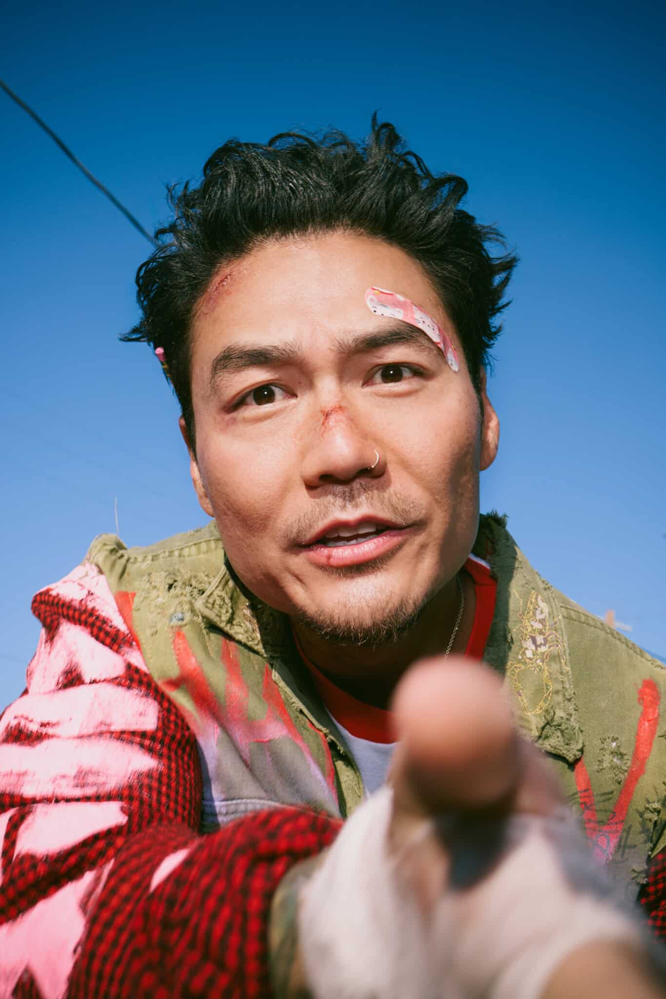 A man with tousled hair and a cut on his eyebrow looks directly at the camera, pointing forward. He wears a colorful, paint-splattered jacket, and the sky is bright blue in the background.