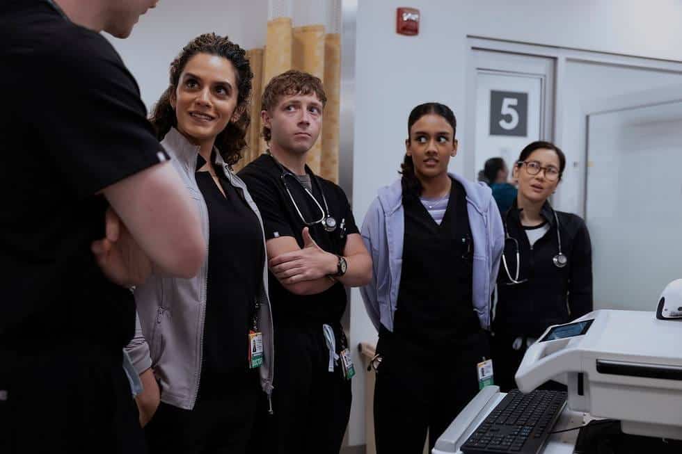 Five medical professionals in scrubs and stethoscopes stand in a hospital hallway, gathered around a medical cart, engaged in conversation and appearing attentive.
