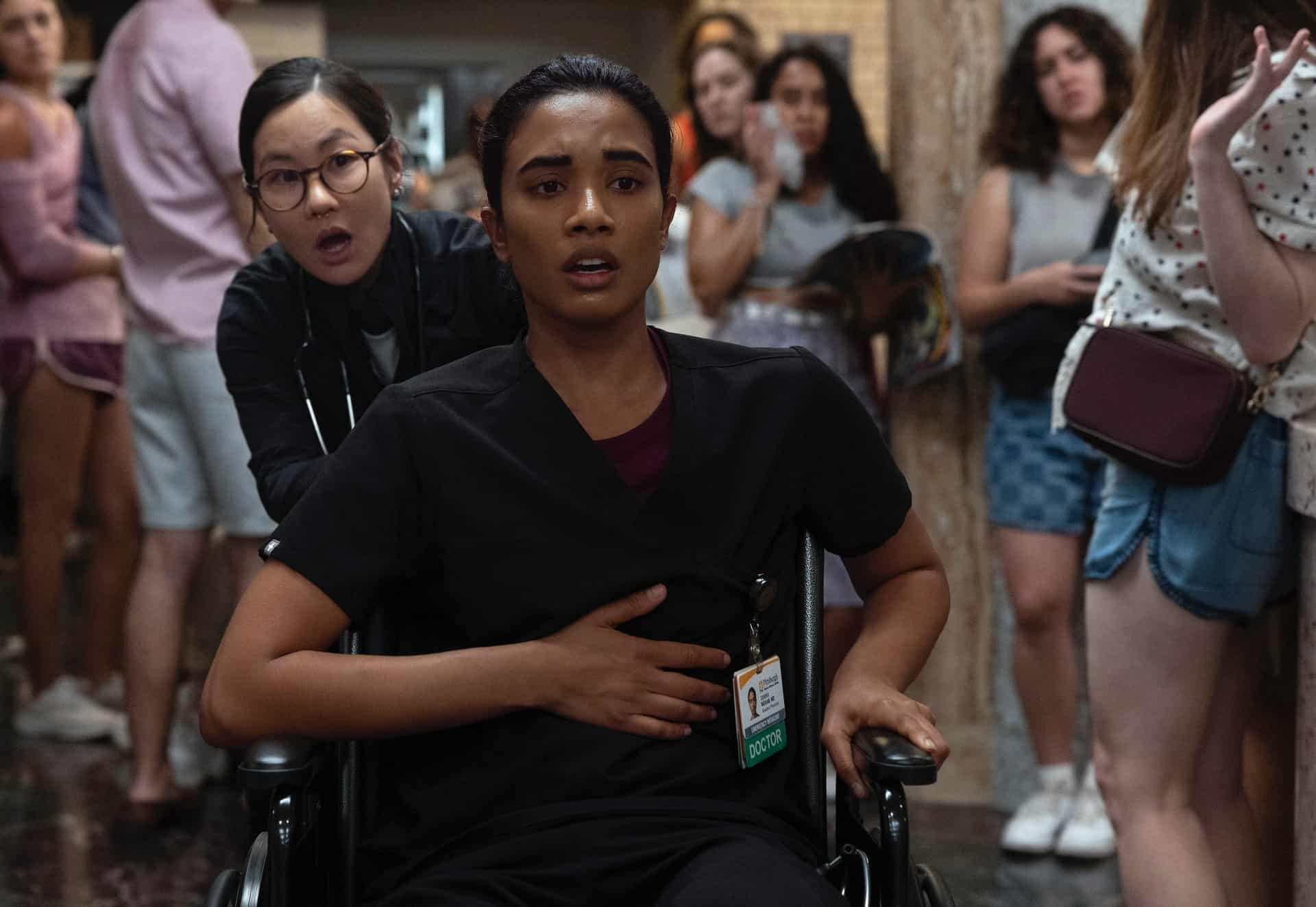 A woman in medical scrubs holds her stomach while sitting in a wheelchair, being pushed by another woman in scrubs. They look concerned. Several people stand in the background watching the scene unfold.