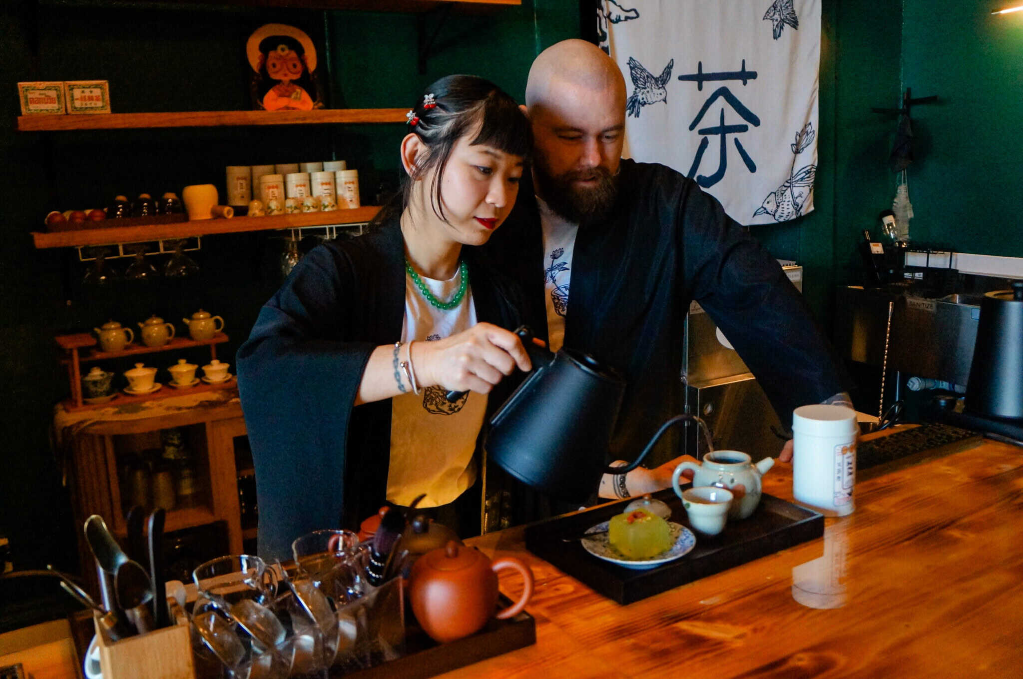 A woman and a man in traditional clothing prepare tea together at a wooden counter in a cozy teahouse, surrounded by teapots, cups, and a dessert. Japanese decor and tea utensils fill the background.