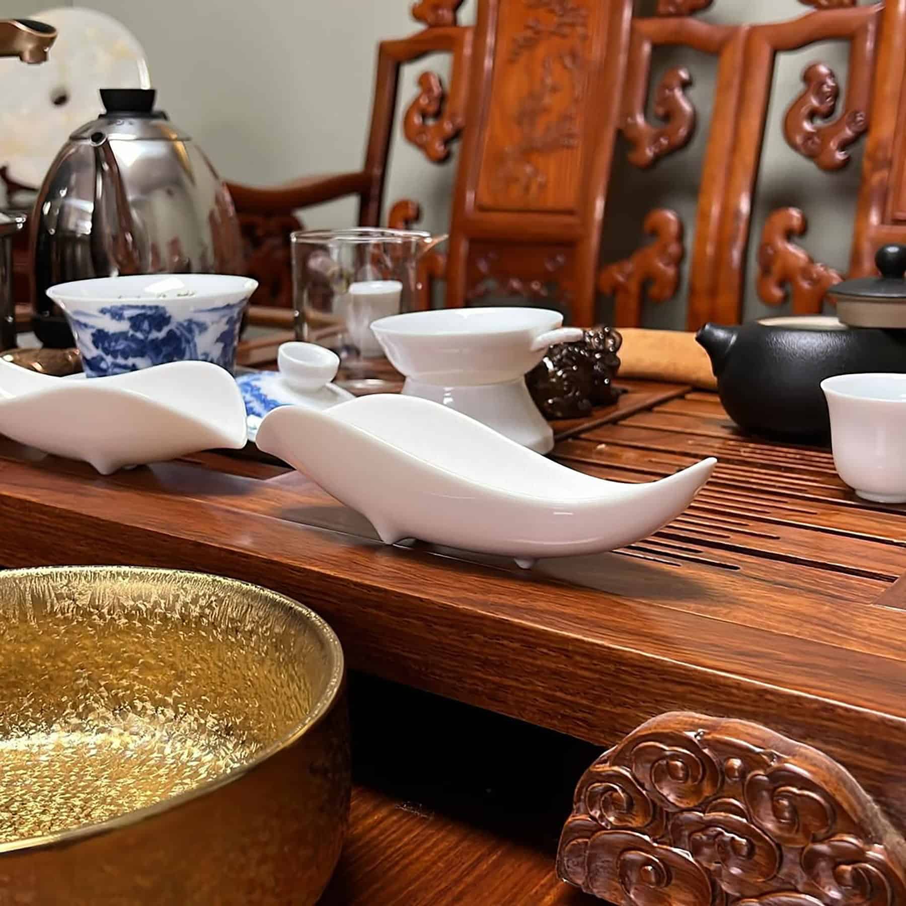 A close-up of a traditional Chinese tea set on a carved wooden table, featuring teapots, cups, a gold bowl, and white porcelain rest shaped like birds, with intricately carved wooden chairs in the background.