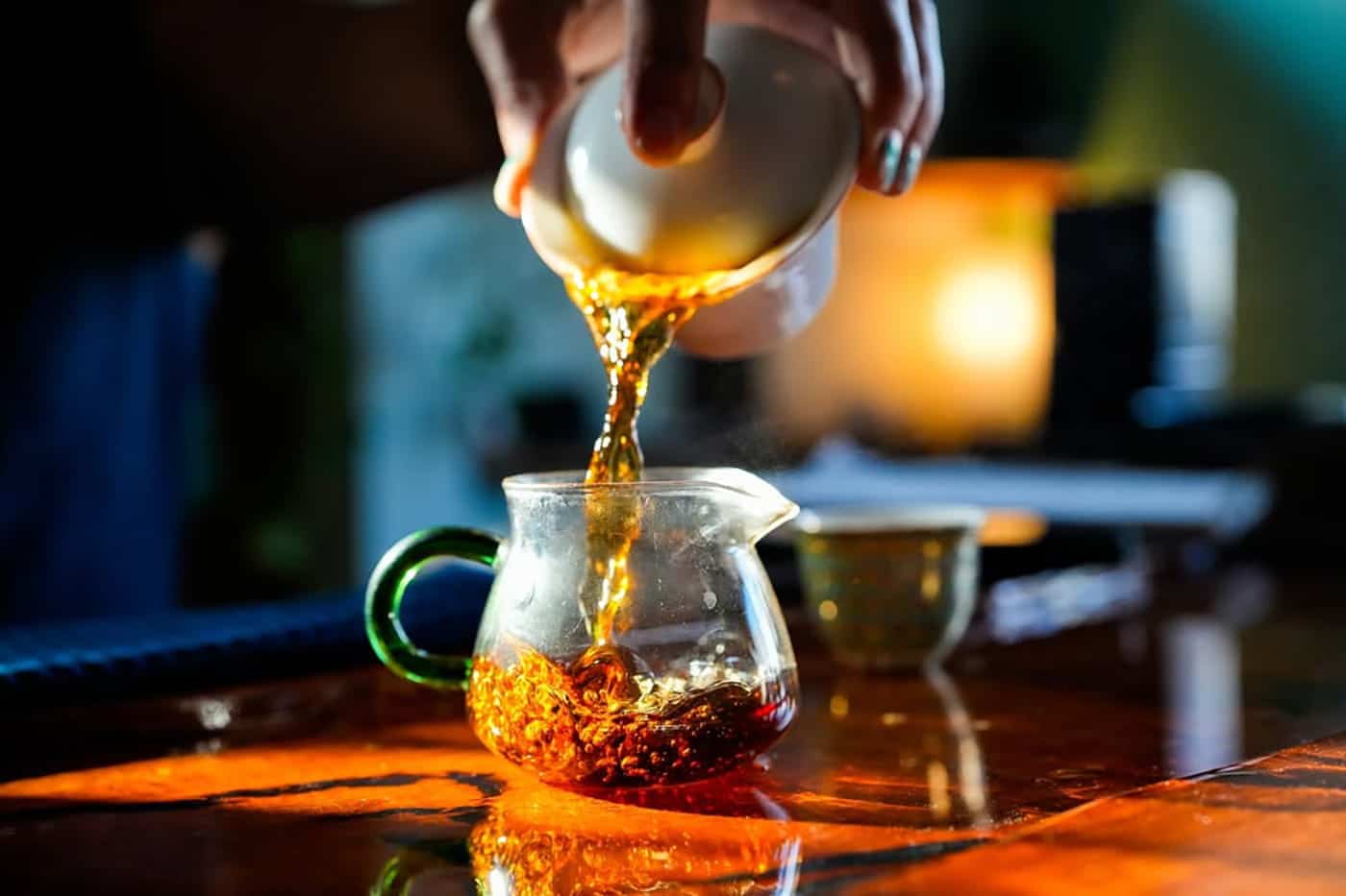 A close-up of tea being poured from a white cup into a glass teapot on a wooden table, with sunlight reflecting off the surface and dried tea leaves visible inside the teapot.