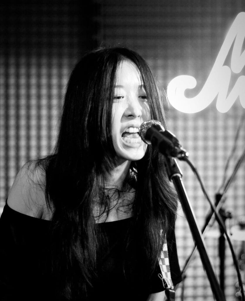A woman with long dark hair passionately sings or screams into a microphone on stage, with a blurred, lit background and a visible drum set behind her. The image is in black and white.