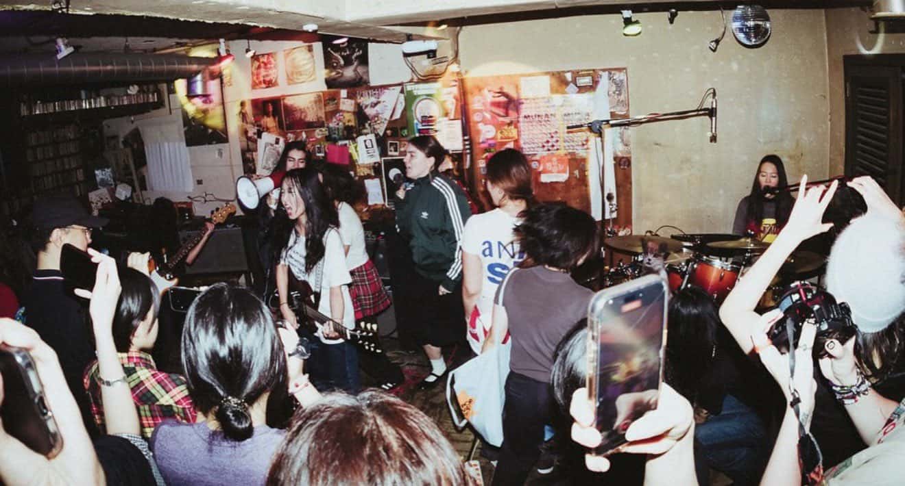 A lively indoor concert scene with a band performing on stage and an enthusiastic crowd. Audience members raise phones and cameras, while colorful posters cover the walls, creating a vibrant, energetic atmosphere.