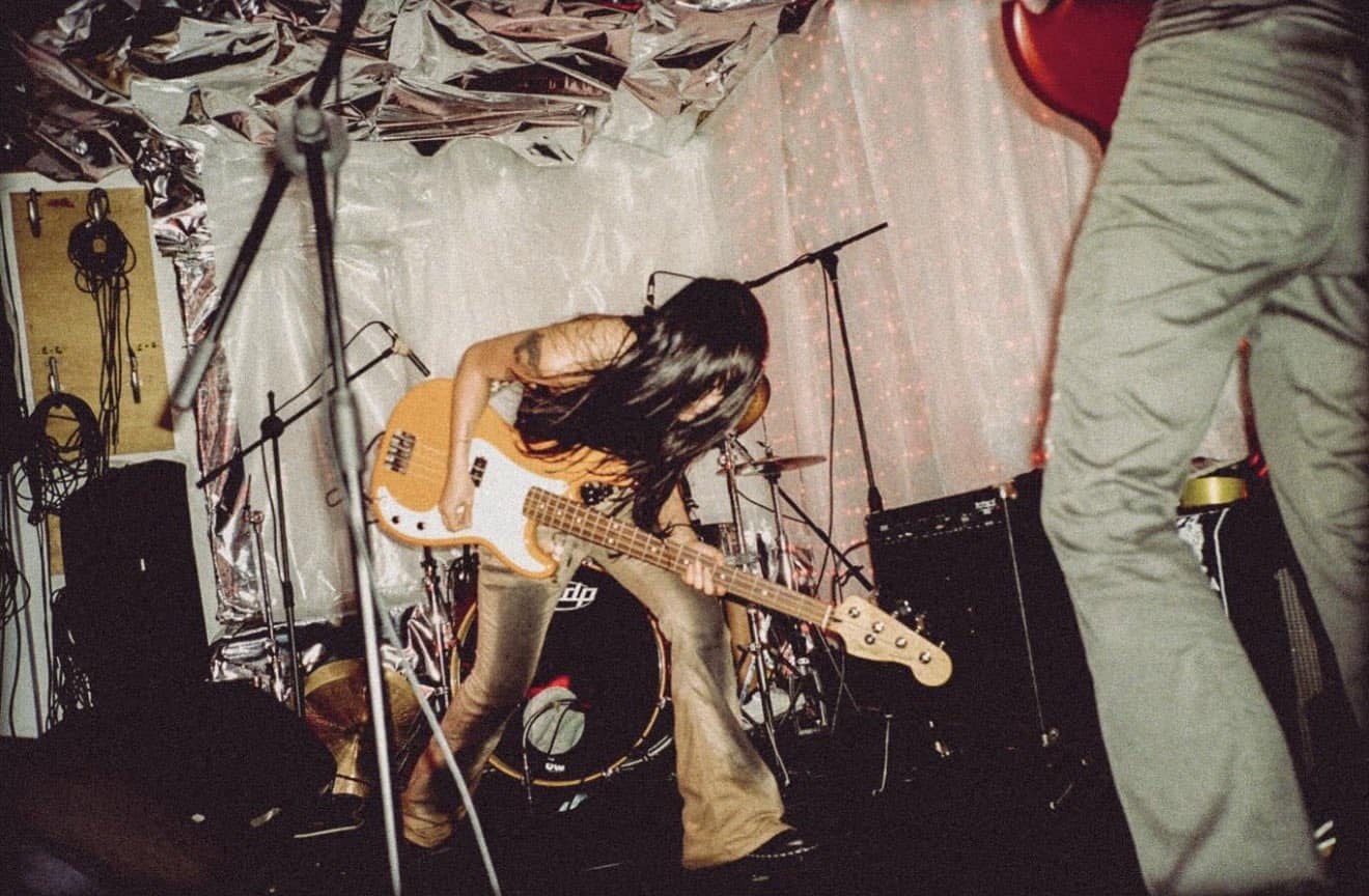 A musician with long hair plays an electric bass guitar energetically on stage, surrounded by drums and amplifiers, with metallic and sheer backdrops and another person partially visible in the foreground.