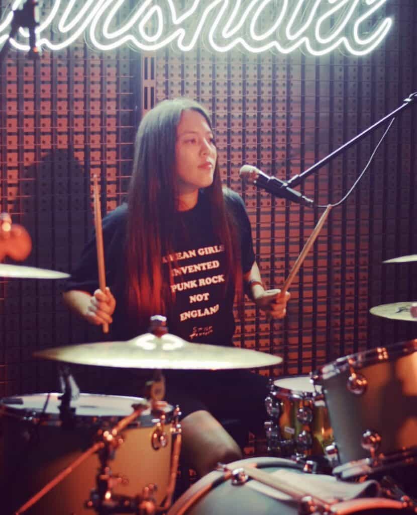 A woman with long hair plays drums under neon lights, wearing a black shirt that reads Asian Girls Invented Punk Rock Not England. She holds drumsticks and looks focused, with a microphone in front of her.