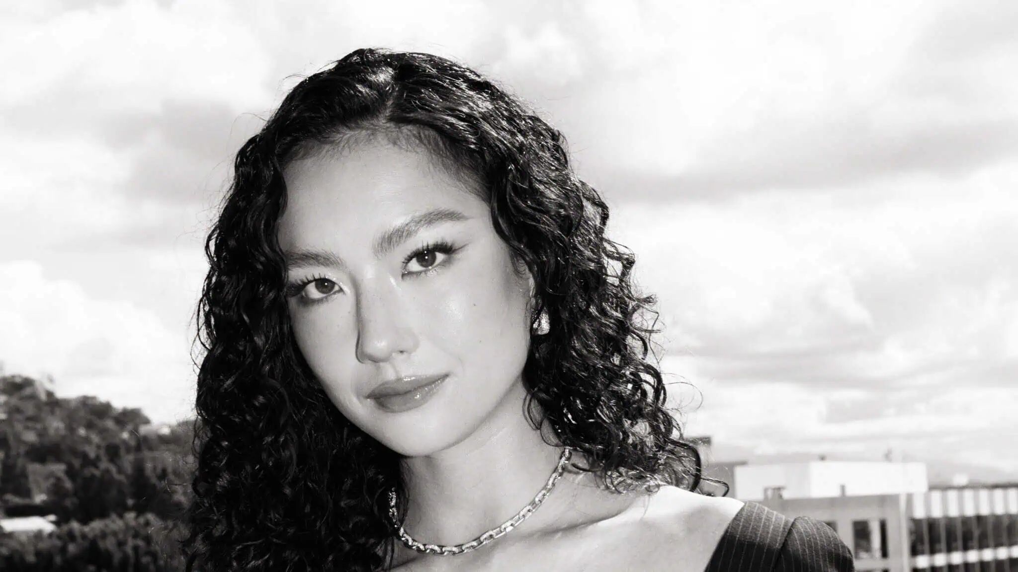 Black and white photo of a woman with long, curly dark hair, wearing a necklace and a dark top, looking at the camera with a neutral expression. The background shows a cloudy sky and some buildings.