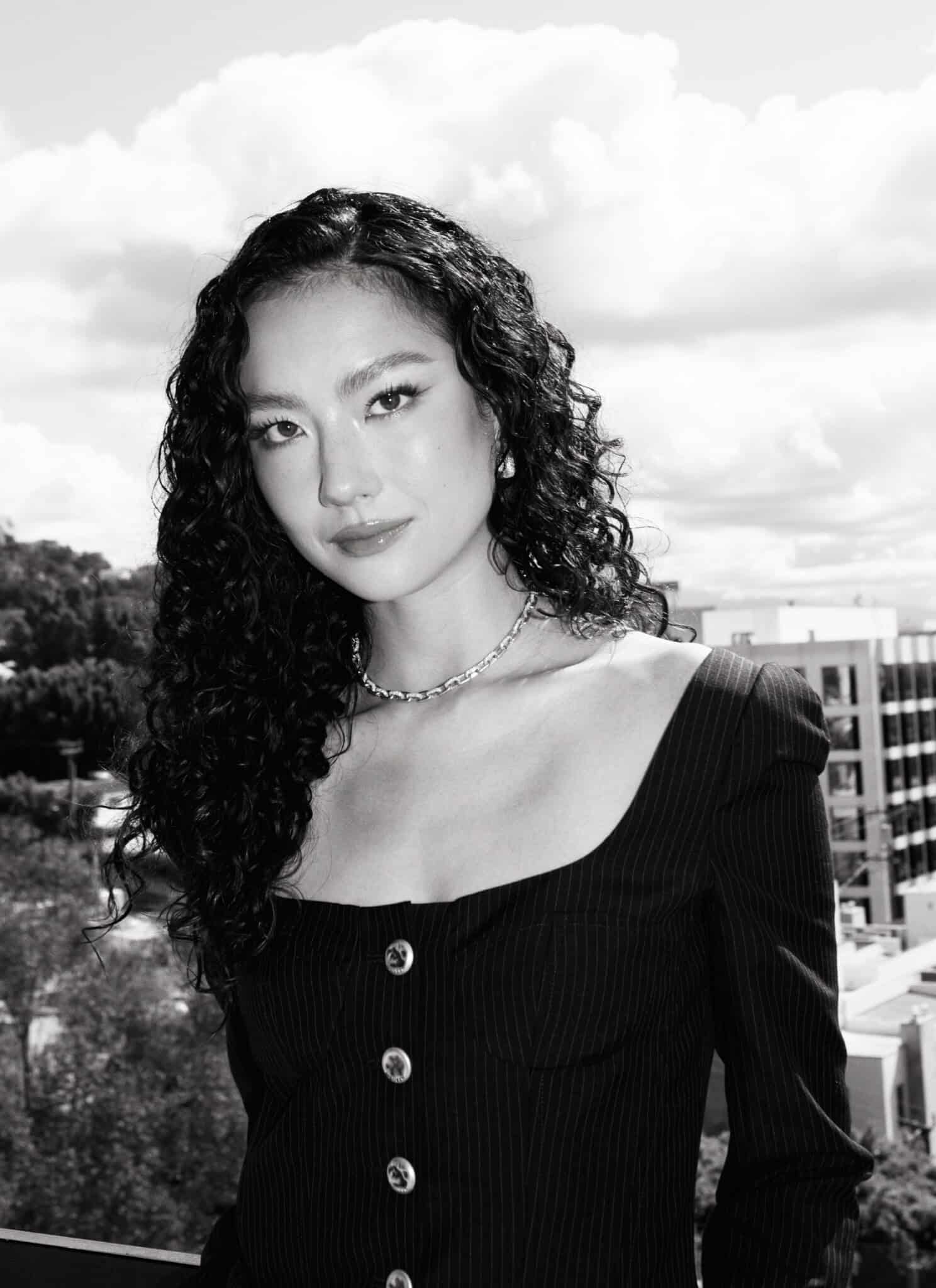 Black and white photo of a woman with long, curly dark hair, wearing a necklace and a dark top, looking at the camera with a neutral expression. The background shows a cloudy sky and some buildings.