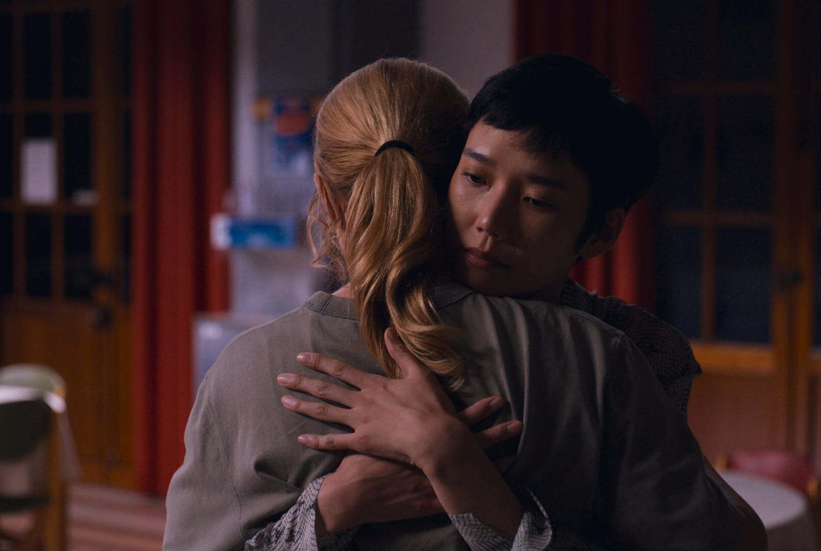 Two people are hugging indoors. The person facing the camera has short dark hair and a thoughtful expression, while the other has long blonde hair seen from behind. The setting appears to be a softly lit room with red curtains.
