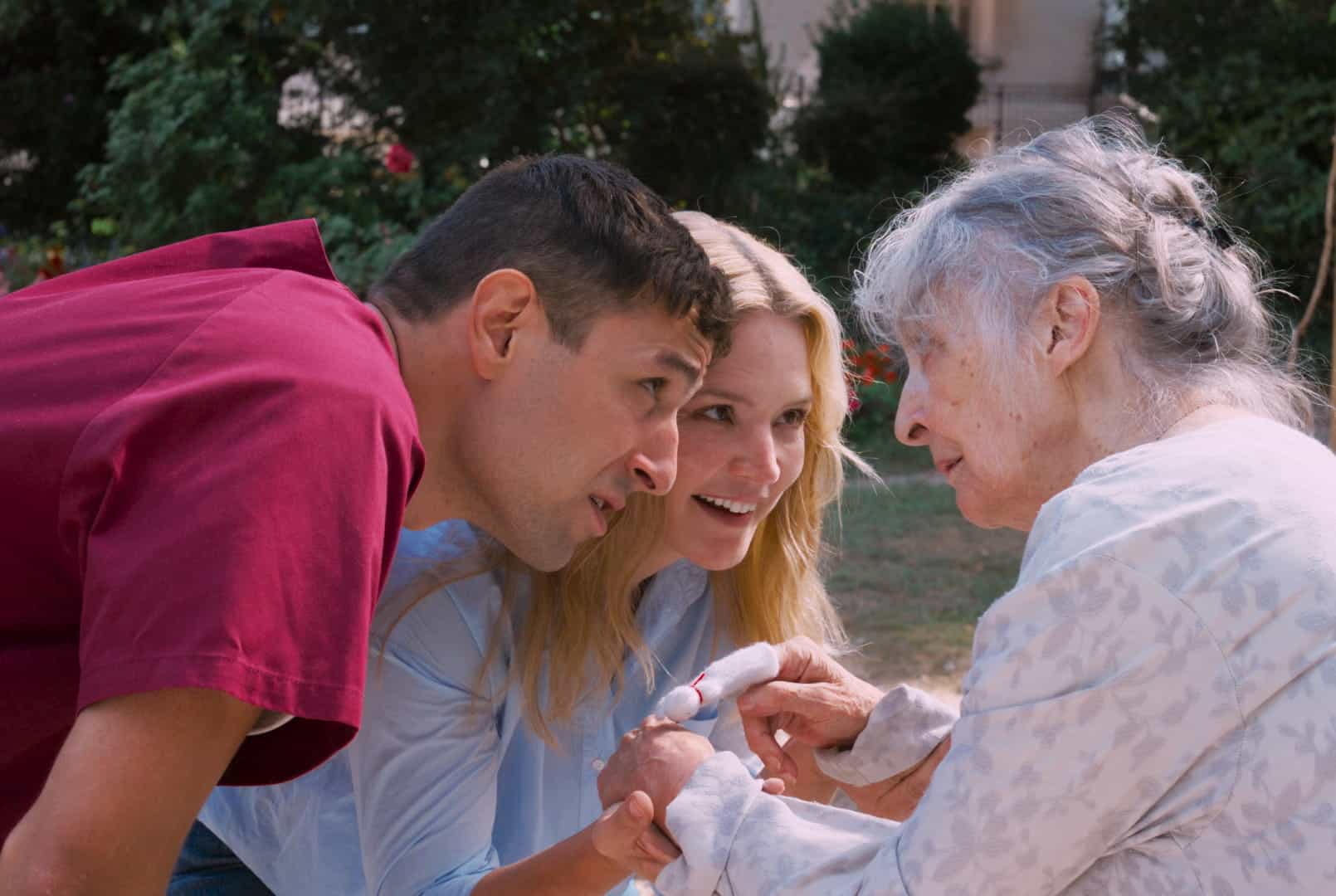 A smiling man and woman kneel down outdoors, gently holding hands and speaking with an elderly woman who appears happy and engaged. Trees and greenery are visible in the background.