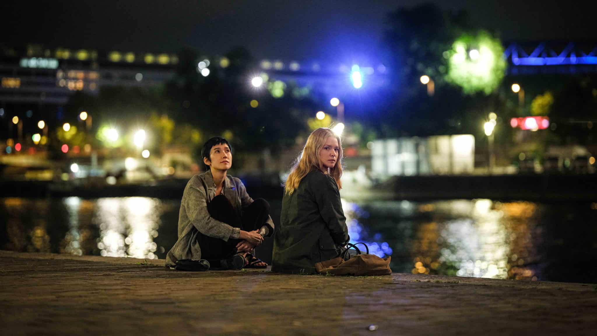 Two people sit on a riverside at night, facing away from each other. City lights and reflections shimmer on the water in the background, with trees and buildings softly illuminated.