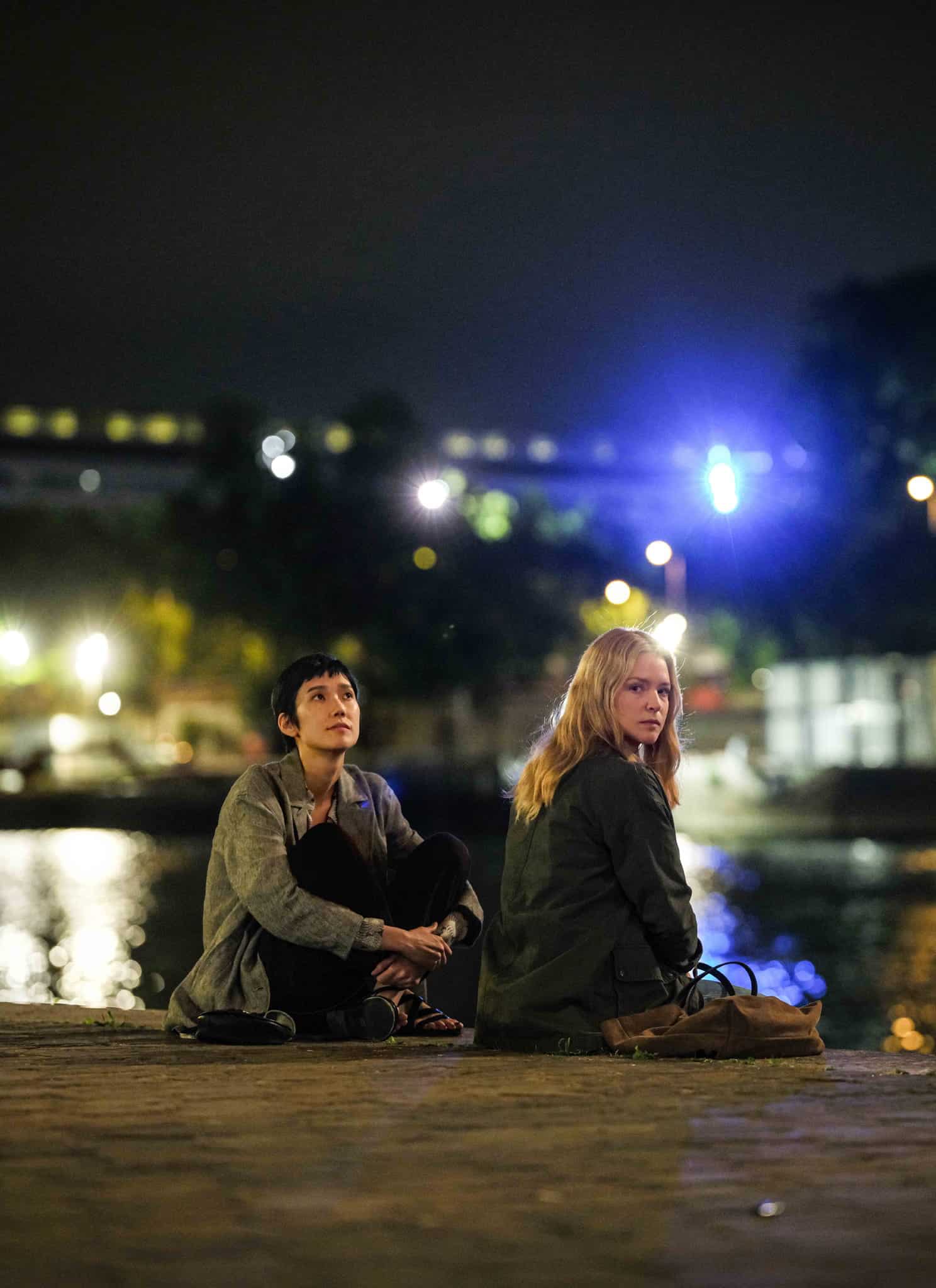 Two people sit on a riverside at night, facing away from each other. City lights and reflections shimmer on the water in the background, with trees and buildings softly illuminated.