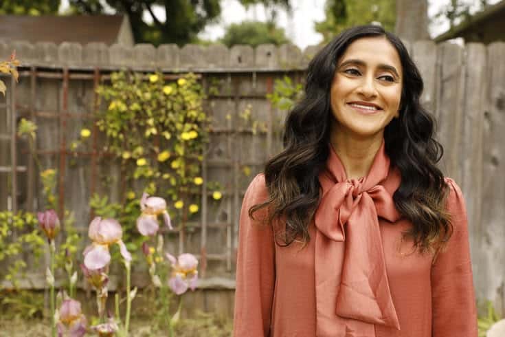 A woman with long, wavy dark hair wearing a rust-colored blouse with a bow stands smiling in a garden with purple flowers and a wooden fence in the background.