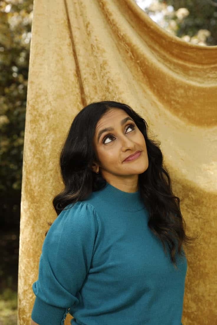 A woman with long dark hair wearing a teal sweater stands in front of a gold velvet backdrop, looking upward with a thoughtful expression. Trees and sunlight are visible in the background.