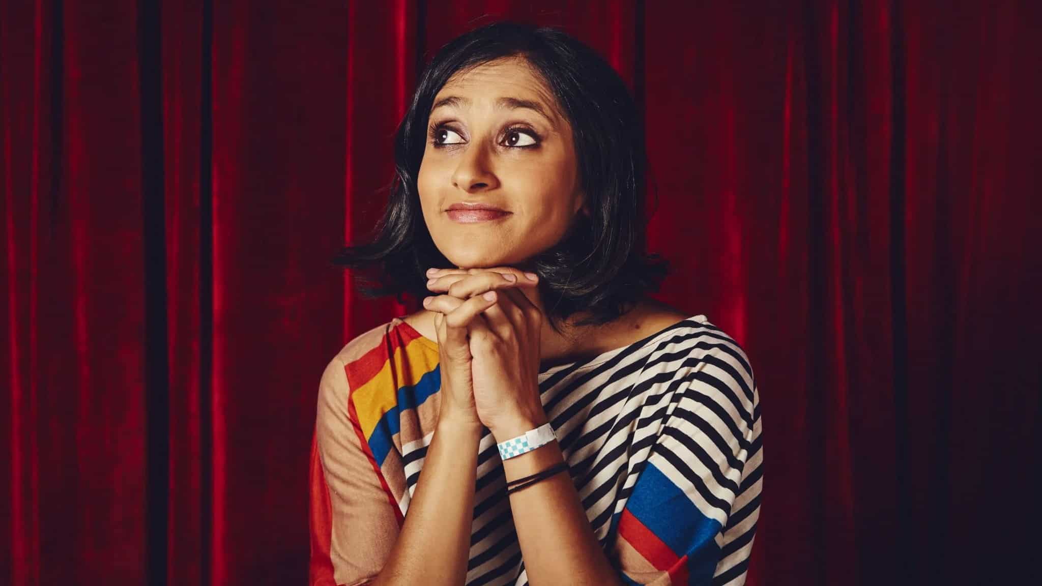 A woman with short dark hair sits in front of a red curtain, smiling slightly and resting her chin on her clasped hands. She wears a striped top with colorful patterns and looks thoughtfully to the side.
