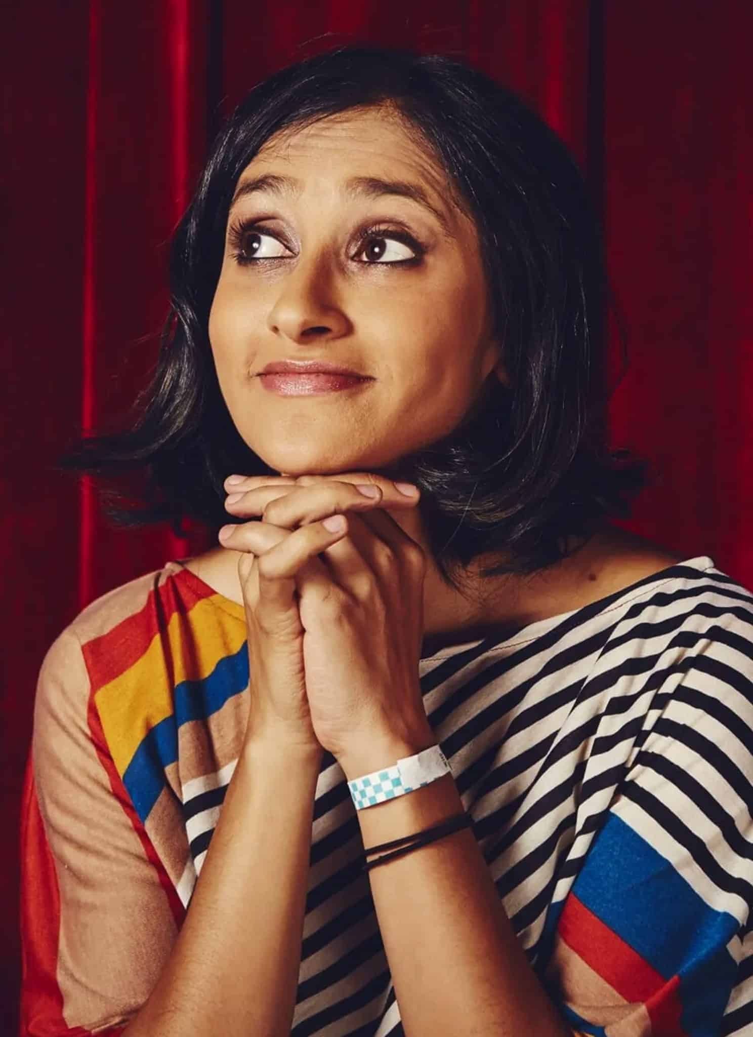 A woman with short dark hair sits in front of a red curtain, smiling slightly and resting her chin on her clasped hands. She wears a striped top with colorful patterns and looks thoughtfully to the side.