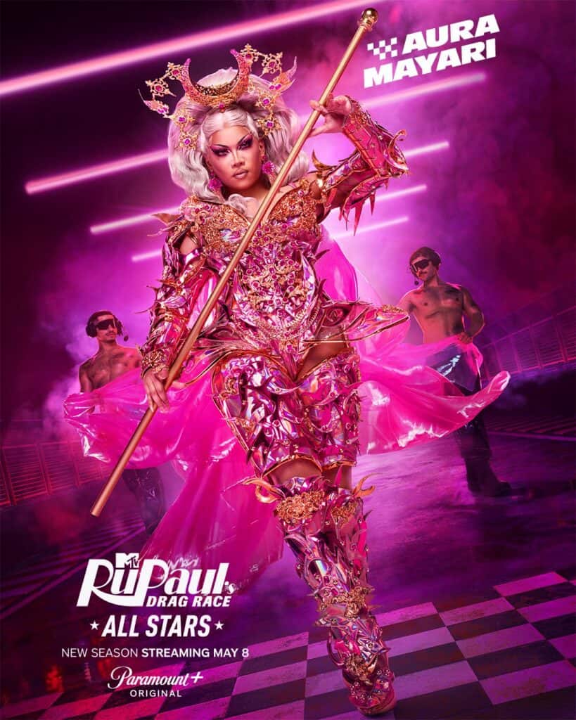 A drag queen dressed in elaborate, pink and gold armor with a crown and scepter poses confidently. Two shirtless dancers stand behind. Text reads Aura Mayari, RuPauls Drag Race All Stars, and Streaming May 8 on Paramount+.