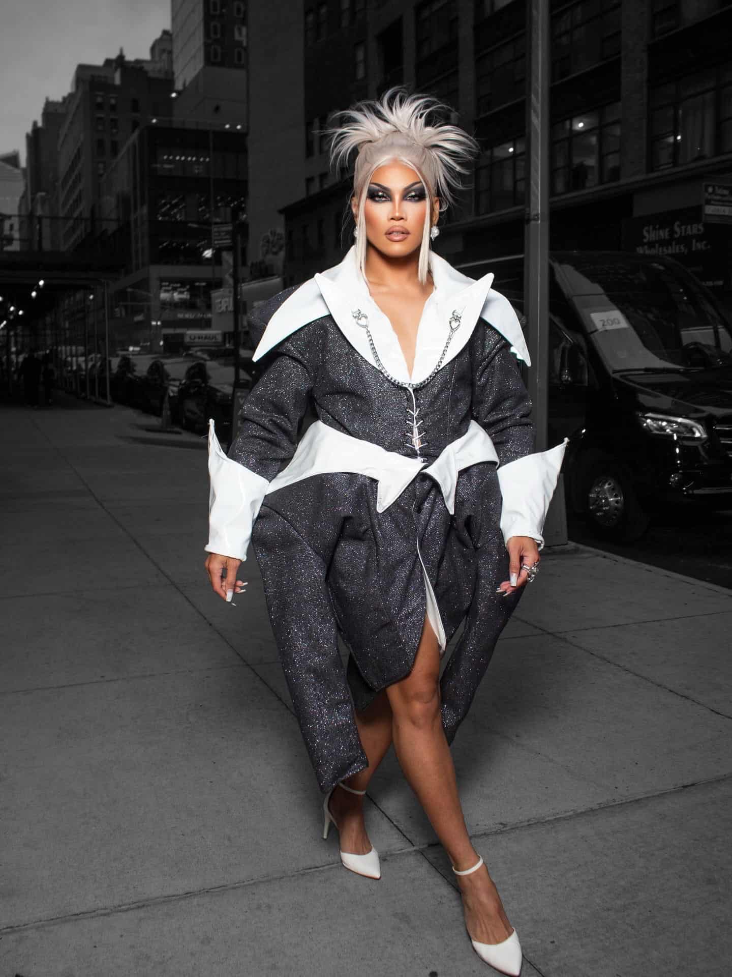 A person with dramatic makeup and spiked silver hair walks confidently on a city sidewalk at night, wearing a shimmering black and white structured coat and white heels. Tall buildings and parked cars are in the background.