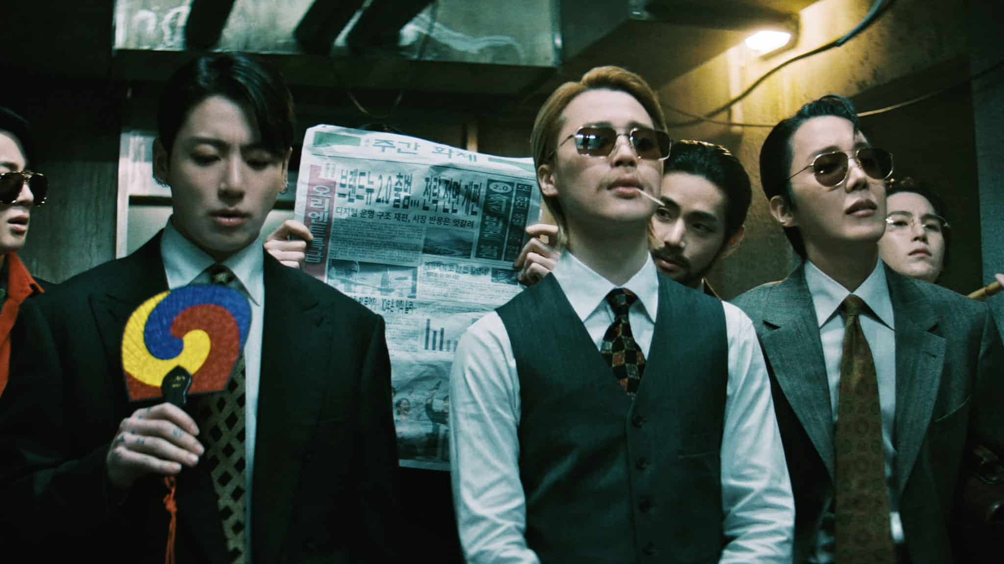 A group of men in suits and sunglasses stand together indoors; one holds a colorful traditional fan and another holds up a Korean newspaper in the background. The scene has a dramatic, vintage atmosphere.