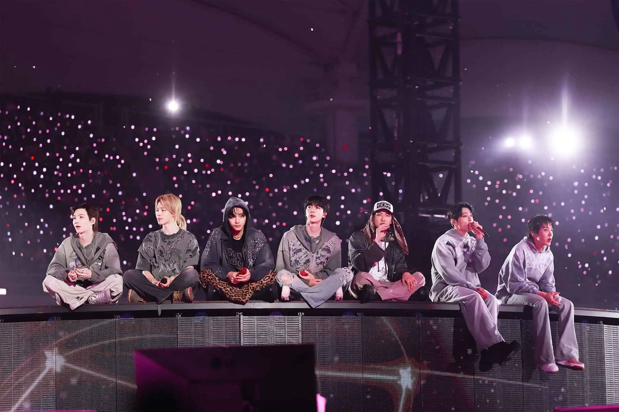 Seven men sit in a row on stage during a concert, wearing casual outfits reminiscent of the BTS Oldboy music video. The background glows with purple and white lights from the audience, creating a vibrant atmosphere.