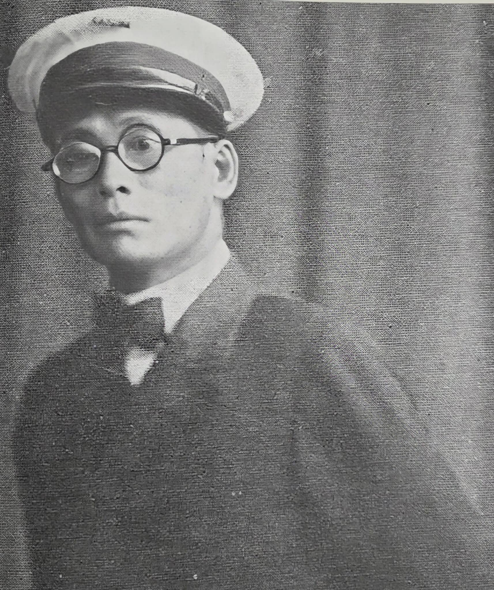 A black-and-white photo of an Asian man wearing round glasses, a peaked cap, a suit, and a bow tie, standing in front of a plain curtain background.