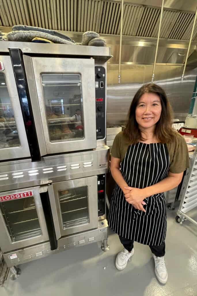 A woman wearing a striped apron stands and smiles next to two large commercial ovens in a professional kitchen. She has long dark hair and is wearing a green t-shirt, black pants, and white sneakers.