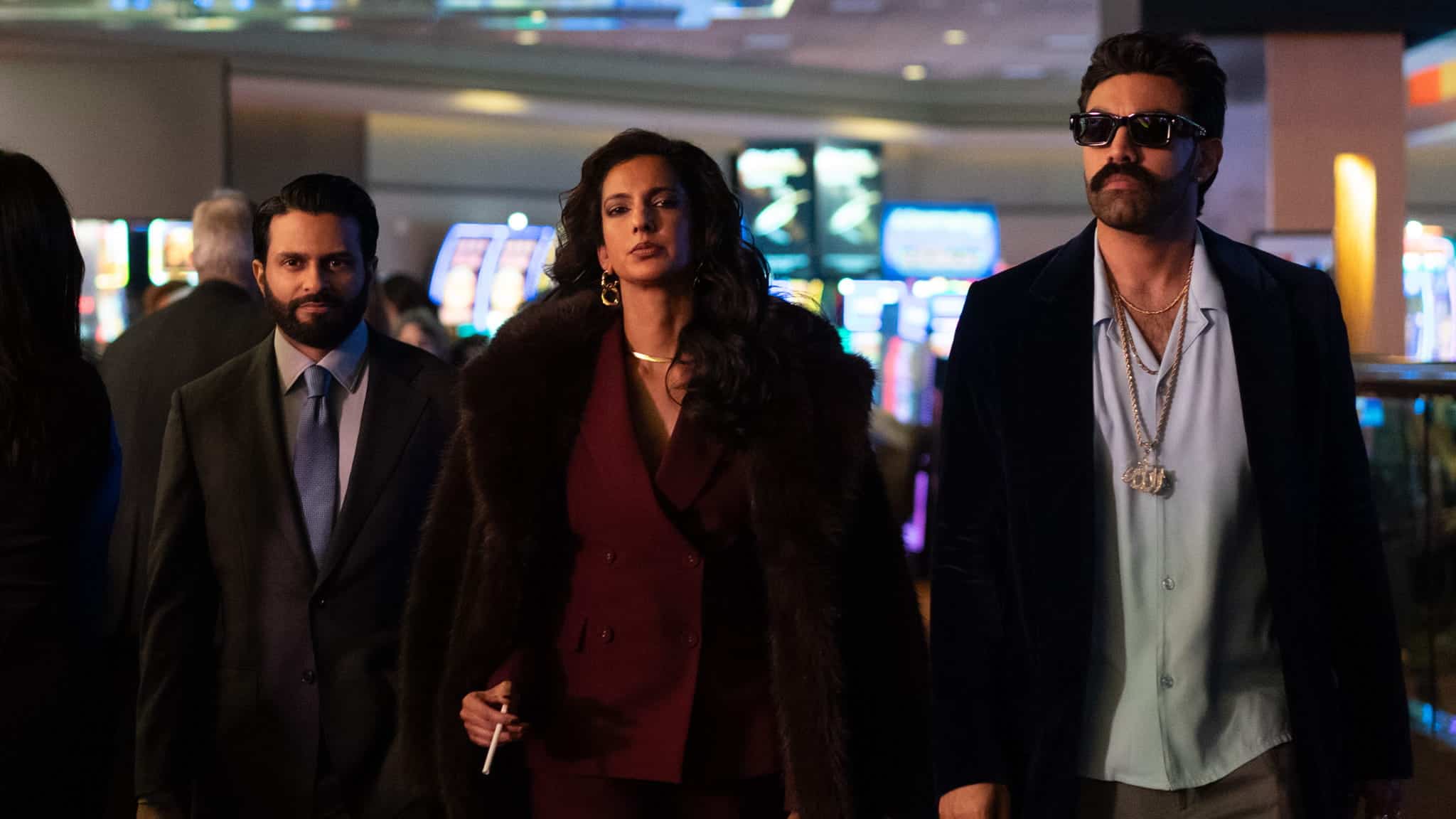Three well-dressed people walk confidently through a casino, with slot machines in the background. The woman in the center wears a fur coat, while the man on the right wears sunglasses and flashy jewelry.
