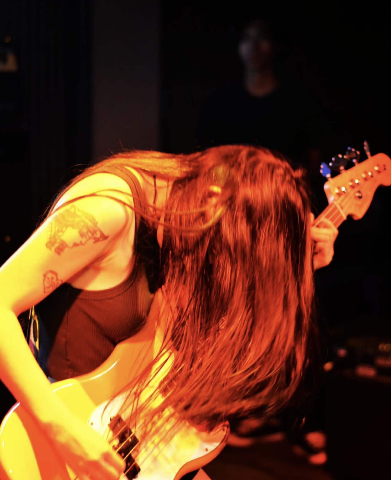 A guitarist with long hair plays an electric guitar passionately on stage, head down with hair covering their face. The lighting is warm and dramatic, highlighting the musicians intensity.