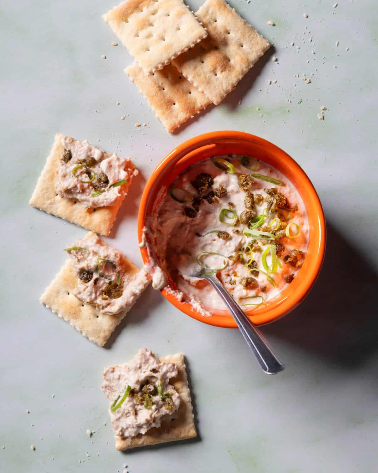 An orange bowl of creamy dip with chopped green onions and bits on top, surrounded by saltine crackers, some spread with the dip. A spoon rests in the bowl. Crumbs are scattered on a light surface.