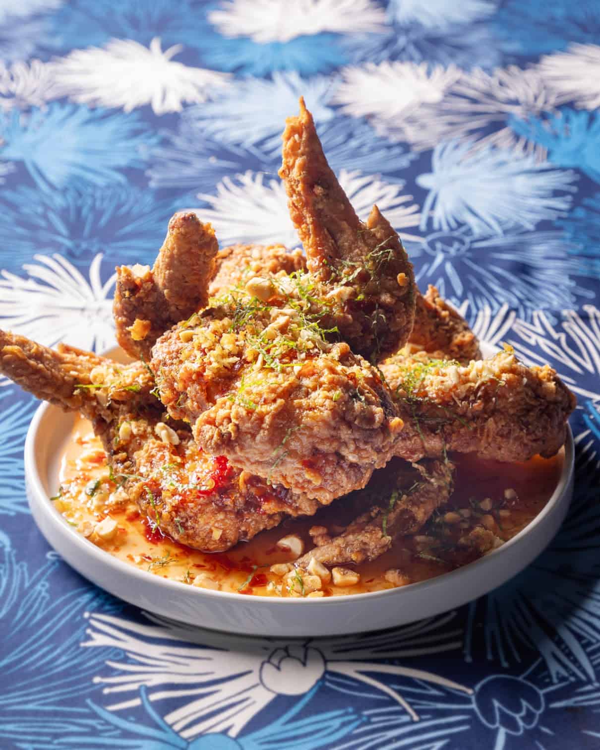 A plate of crispy fried chicken wings garnished with chopped herbs and drizzled with a reddish sauce, served on a dish set on a blue and white patterned tablecloth.