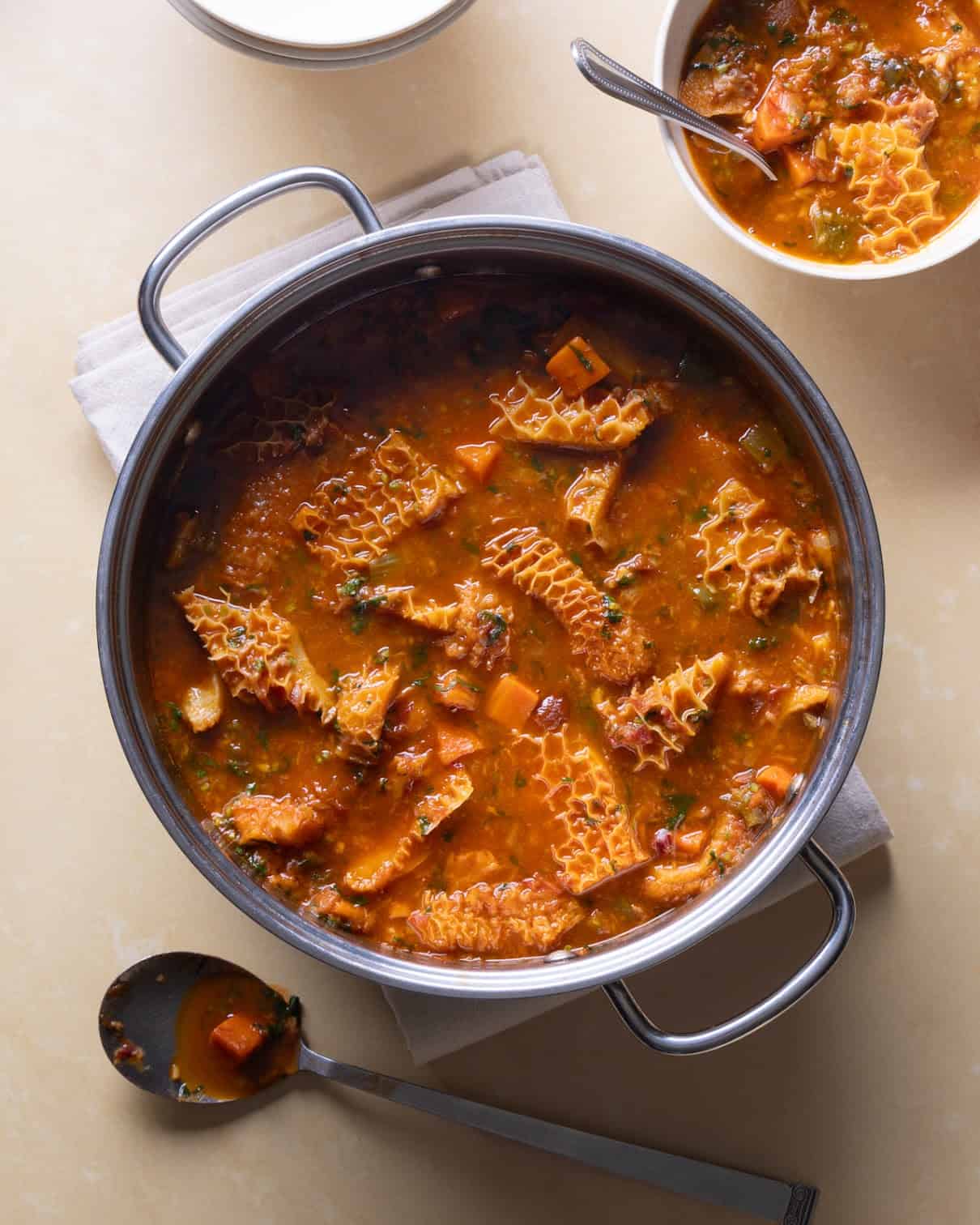 A pot of tripe stew with tomato-based broth, visible honeycomb tripe pieces, carrots, and herbs. A filled bowl and a spoon rest nearby on a beige surface.
