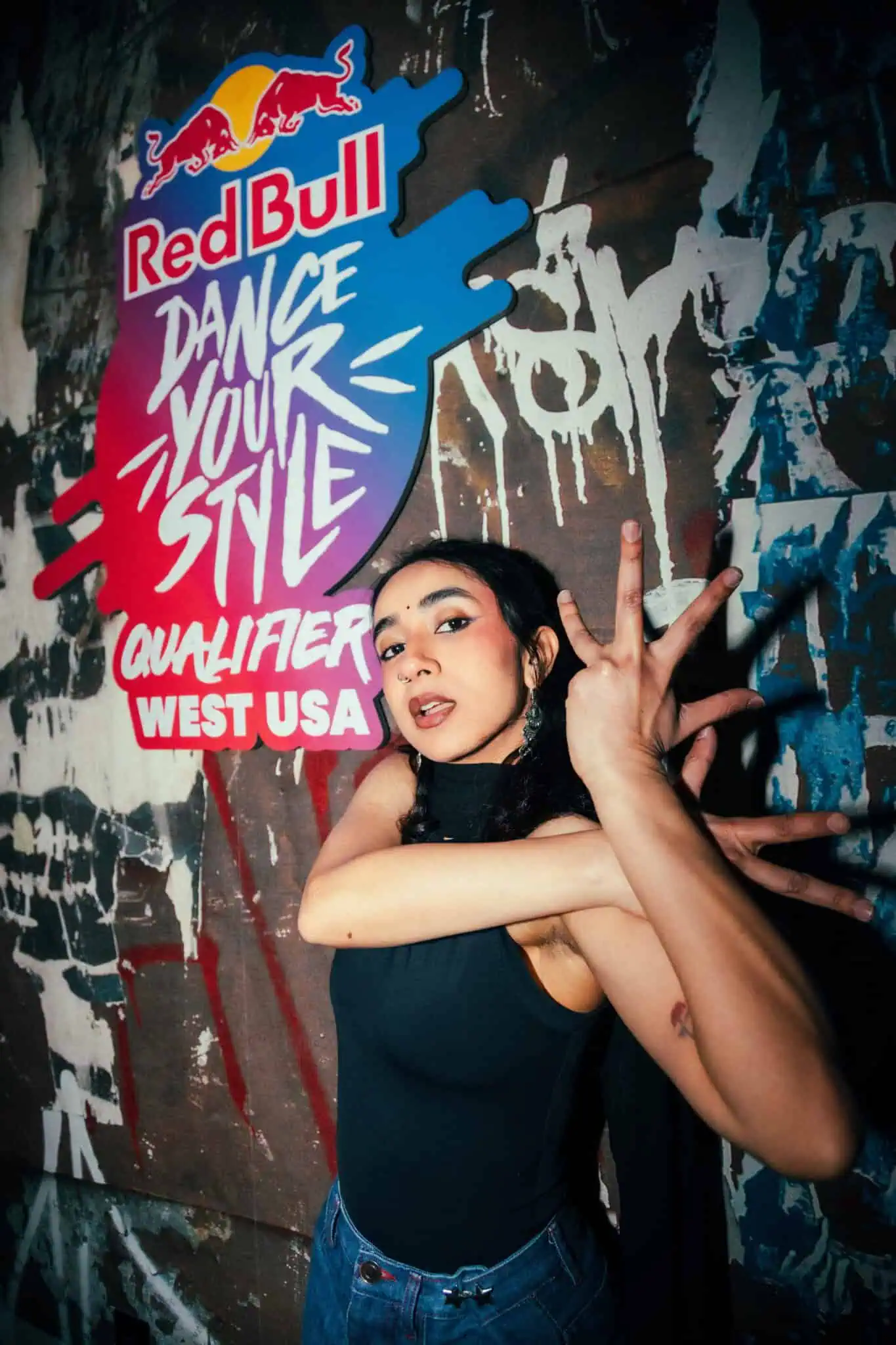 A dancer poses with expressive hand movements in front of a colorful wall sign that reads “Red Bull Dance Your Style Qualifier West USA.” The scene has an urban, graffiti-inspired background.