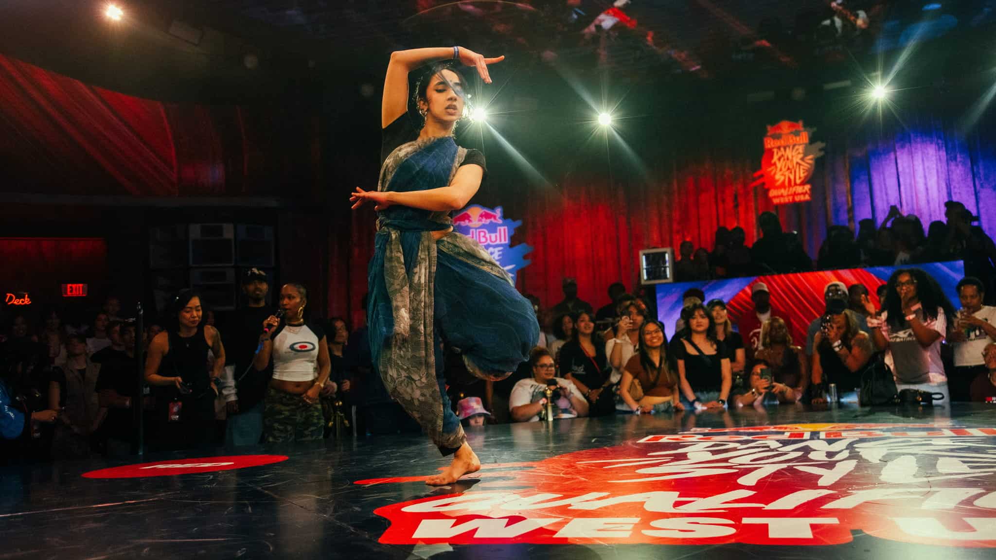 A dancer in traditional attire performs on stage at a dance competition, surrounded by a cheering crowd under colorful lights and vibrant decorations.