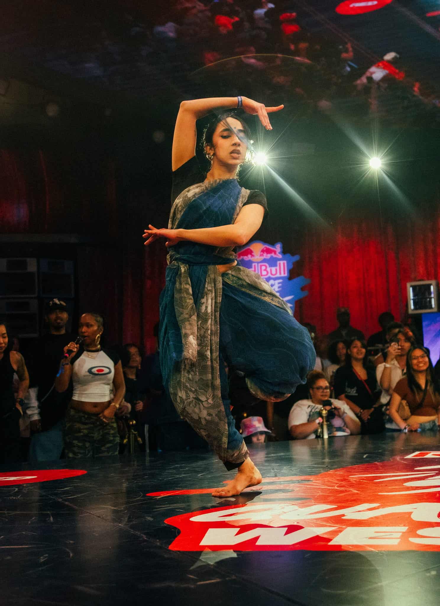 A dancer in traditional attire performs on stage at a dance competition, surrounded by a cheering crowd under colorful lights and vibrant decorations.