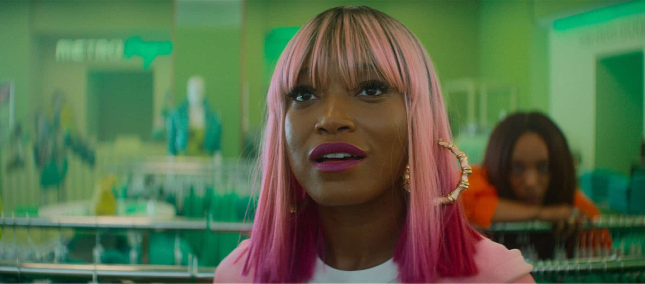 A woman with straight pink hair, blunt bangs, and large gold hoop earrings looks upward and smiles in a brightly lit laundromat. Another person sits in the background, leaning on a counter. The scene has a green tint.